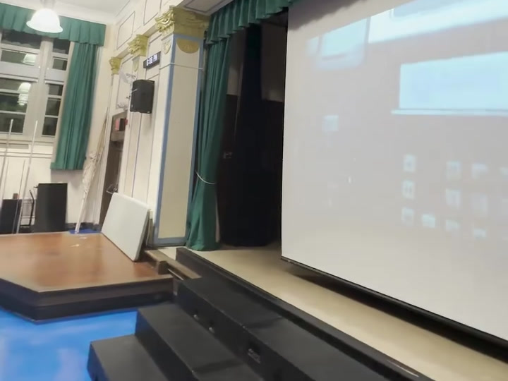 Auditorium stage with an active projection screen, green curtains and a wall-mounted speaker.